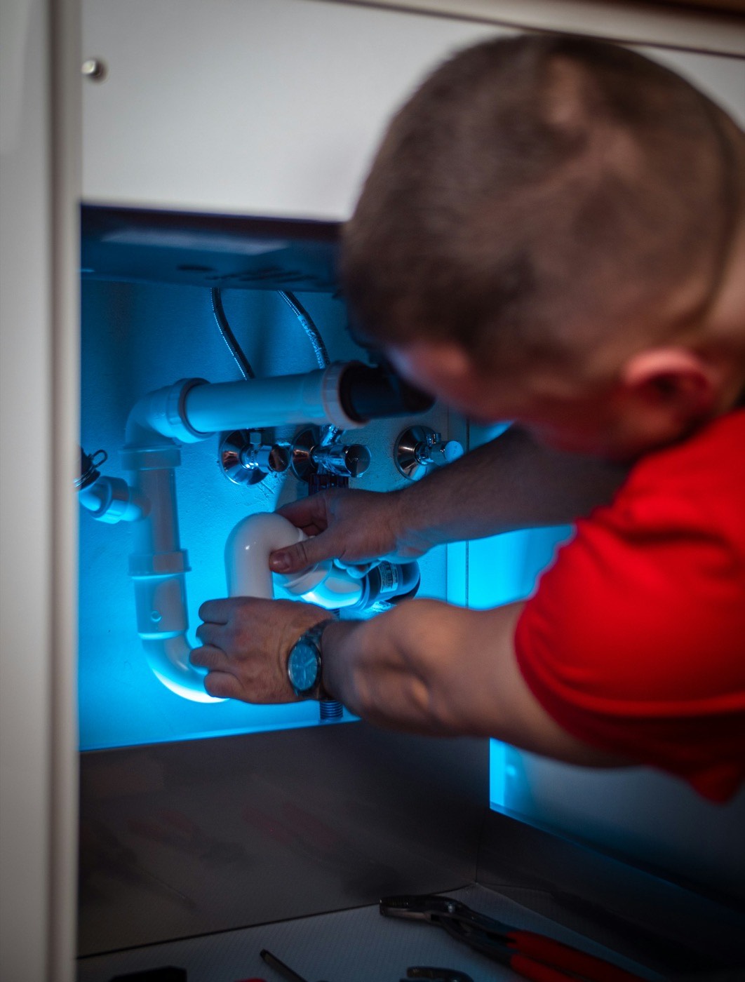 A Hartley plumber tightening a trap under a kitchen sink by work light.